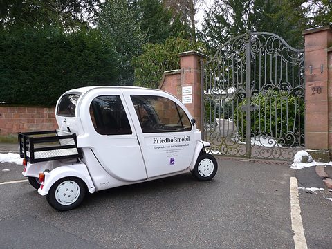 White four-seat electric car lettered Friedhofsmobil with a rear rack, parked by iron cemetery gates. Opens a larger view