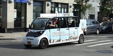 Long white six-seat electric shuttle with an advertising wrap crossing a street junction, shops behind. Opens a larger view