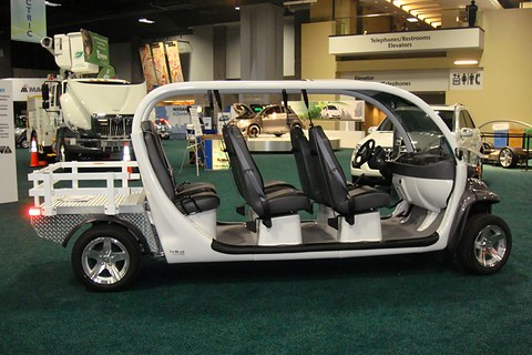 Side view of a long white open electric car with three rows of black seats and a rear deck on green carpet.