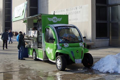 Green electric car with a food counter on its rear beside a stone museum wall and a heap of snow. Opens a larger view