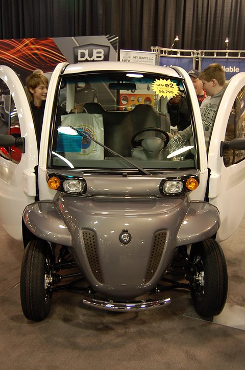 Front of a grey and white electric car with both doors open, a twin-bar grille and round lamps on carpet. Opens a larger view