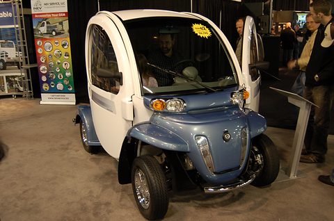Blue and white two-seat electric car with round lamps and doors open on a show floor, visitors around it.