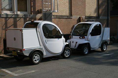 Two white two-seat electric utility vehicles with box beds parked beside a brick building. Opens a larger view