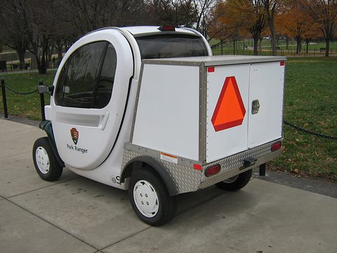 Rear of a white electric utility vehicle with a box body carrying an orange slow-vehicle triangle on paving. Opens a larger view
