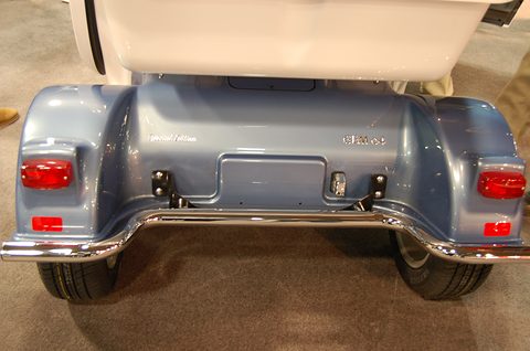 Rear of a blue electric car with red lamps, a chrome bumper bar and a white upper body on a show carpet. Opens a larger view