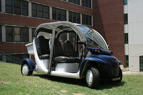 Dark blue and white open four-seat electric car with a canvas roof parked on grass before a brick building. Opens a larger view