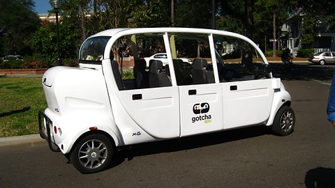Long white six-seat electric car with doors and ‘gotcha’ lettering parked on a campus road under trees. Opens a larger view