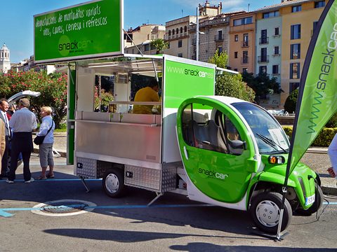 Green electric car towing a steel snack counter with green banners on a square, people standing by. Opens a larger view