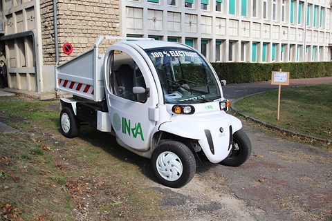 White two-seat electric utility vehicle with INRA lettering and a tipper body on a gravel path by buildings. Opens a larger view