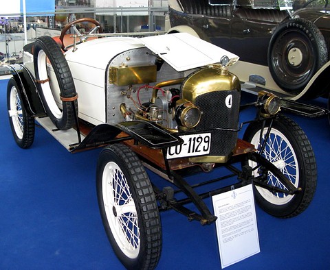 A white two-seater with a brass radiator, open bonnet showing the engine, spare wheel and white wire wheels on blue carpet. Opens a larger view