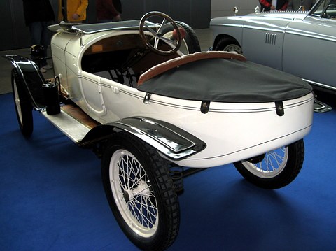 Rear view of a white two-seater with a black tonneau cover on its rounded tail and white wire wheels on blue carpet. Opens a larger view