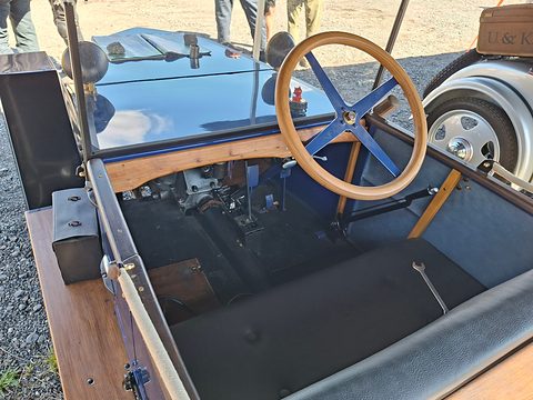Looking down into a small open car: a wooden-rimmed wheel with blue spokes, pedals, gear lever and a black bench seat. Opens a larger view