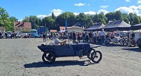 A small blue open car with spoked wheels and a lone driver crossing a gravel yard, a crowd and gazebos behind. Opens a larger view