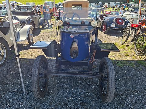 Front view of a tiny blue car with two lamps, a narrow body between flat wings and wire wheels, parked on gravel. Opens a larger view