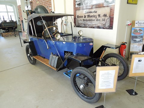 A small blue open car with its hood up, wooden running boards and wire wheels, information boards and a sign on the wall behind. Opens a larger view