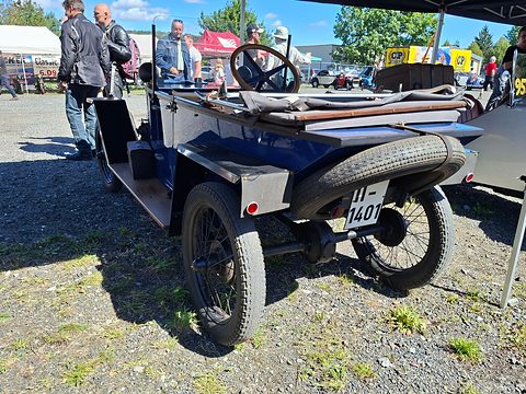 Rear three-quarter view of a small blue open car with a spare tyre on its tail and wire wheels, visitors behind. Opens a larger view
