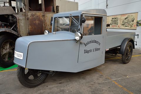 A grey three-wheeled van with a rounded nose, single front lamp and open cargo bed, lettered Kolonialwaren Nägele & Söhne.