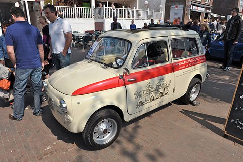 Cream small estate car with a red stripe and cartoon signwriting on brick paving, people beside it. Opens a larger view