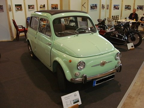 Mint-green small estate car on a dark carpet in a show hall, motorcycles and tables behind. Opens a larger view