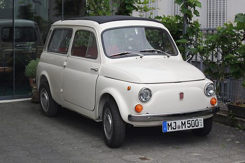 Front of a white small estate car with round headlamps parked on paving beside a hedge. Opens a larger view