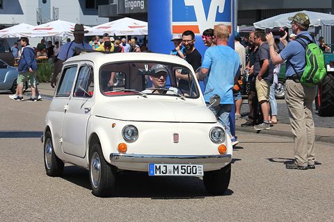 White small estate car driving slowly past spectators at a show, its driver at the wheel. Opens a larger view