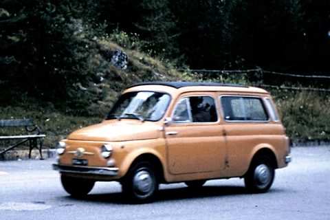 Ochre small estate car with a black fabric roof parked on a mountain road, a wooden bench and pines behind. Opens a larger view