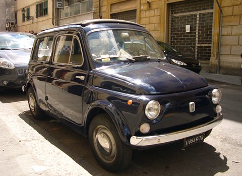 Dark navy small estate with chrome bumpers and round headlamps, parked on a pavement before shuttered shops. Opens a larger view
