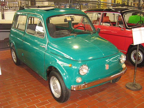Teal small estate car on a red brick floor in a museum, a red car behind. Opens a larger view