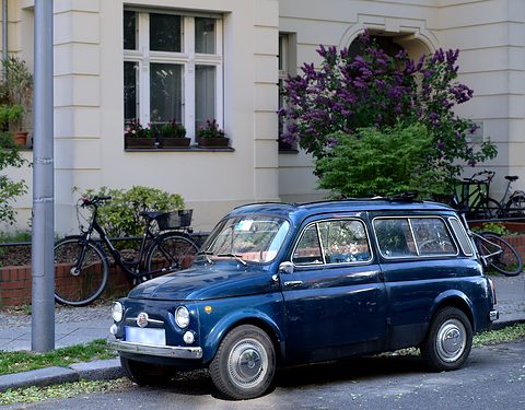 Dark blue small estate car parked at a kerb before a pale house with bicycles and a lilac bush. Opens a larger view