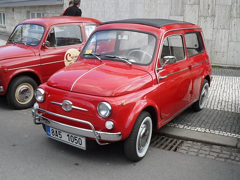 Red small estate car with white-wall tyres parked on cobbles beside another red car. Opens a larger view