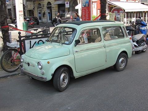 Mint-green small estate with round headlamps, parked on a street beside bicycles and scooters. Opens a larger view