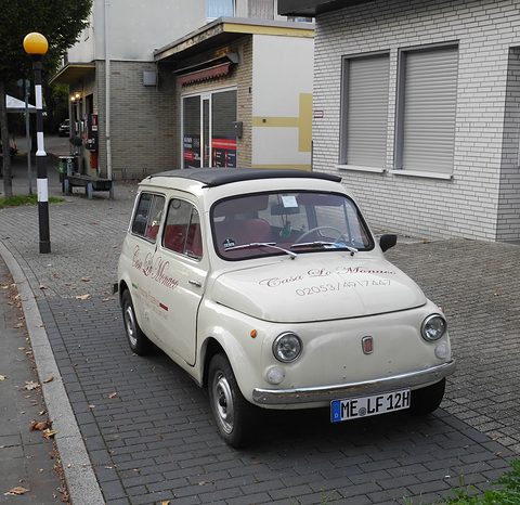 Cream small estate car with script lettering on its bonnet and doors, parked on block paving beside a shop. Opens a larger view