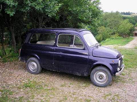 Dark blue small estate car on gravel under green trees, a field behind. Opens a larger view