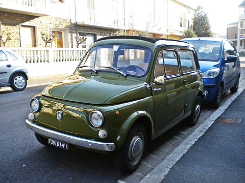 Olive-green small estate car parked at a kerb on a sunny street beside a blue car. Opens a larger view