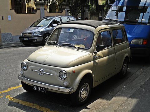 Beige small estate car with chrome bumpers parked by yellow road markings, vans and buildings behind. Opens a larger view