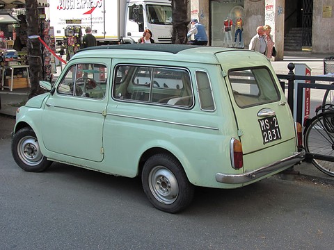 Mint-green small estate car from the rear right, with a black Italian plate, people walking past shops behind. Opens a larger view