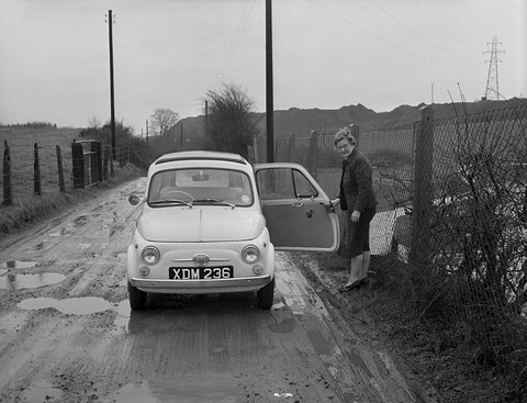 A small pale car on a puddled lane by a wire fence, a woman in a coat standing at its open door, pylons behind. Opens a larger view