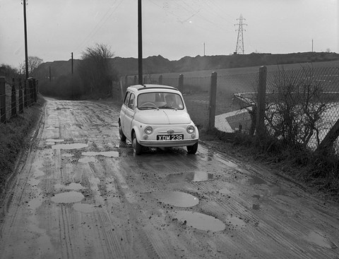 A small white estate car drives along a puddled country lane between fences, pylons behind. Opens a larger view