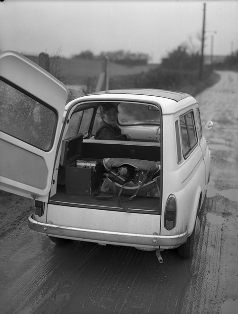 Rear of a small white estate car with its side-hinged back door open, a man at the wheel, on a wet lane.
