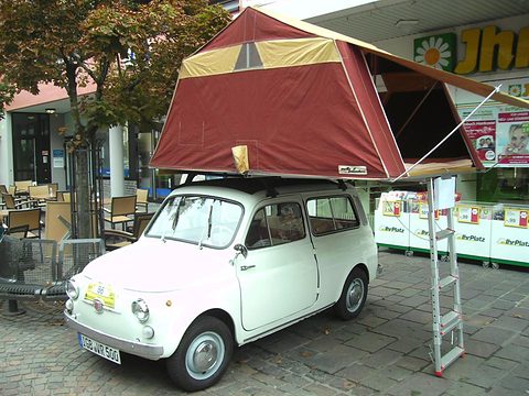 White small estate car with a large red and cream tent on its roof and a ladder to the ground. Opens a larger view