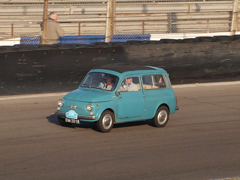 Turquoise small estate car on a race track with a driver and passenger, a spectator above the wall. Opens a larger view