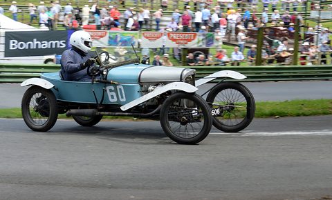 A helmeted driver in a pale blue cyclecar numbered 60 on a tarmac hill, spectators behind barriers. Opens a larger view