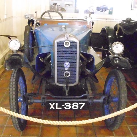 Front view of a blue cyclecar with a mesh radiator, two headlamps and number plate XL-387 behind a rope. Opens a larger view