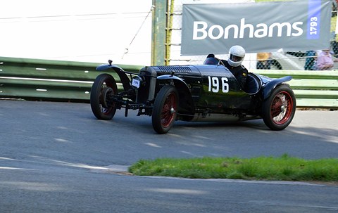 A dark racing special with red wheels rounds a tarmac bend under a Bonhams banner. Opens a larger view