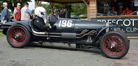 A helmeted driver in a long dark racing special with red wire wheels and exposed exhausts, spectators behind. Opens a larger view