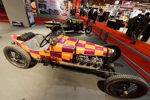 A long racing car with orange and purple chequered bodywork and an exposed aero engine on a grey stand. Opens a larger view