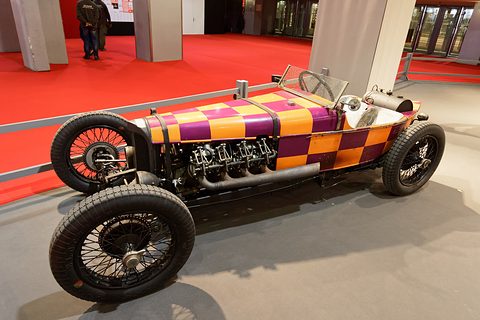 Side view of a racing car with chequered bodywork and an exposed multi-cylinder engine along its flank. Opens a larger view