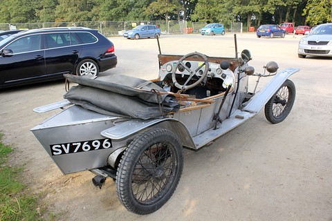 A grey vintage cyclecar with a folded hood on its tail, parked on gravel with modern cars behind. Opens a larger view