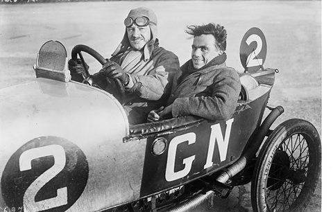 Two men side by side in a pale racing cyclecar lettered GN with a big number 2 on its flank, on a concrete track.