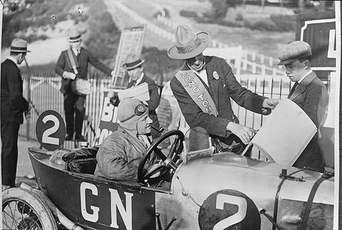 A goggled driver in a pale racing cyclecar lettered GN and numbered 2, a man in a sash and cowboy hat handing him a paper. Opens a larger view